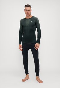 Zeleni sportski top s dugim rukavima i teksturiranim uzorkom, uparen s uskim crnim tajicama. Čini se da je materijal rastezljiv i prozračan.
