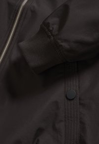 Tumepruun jakk, millel on hõbedane lukk, soonikkoes mansetid ja must snap-nupp. Kangal on sile tekstuur koos minimaalsete õmbluse detailidega.