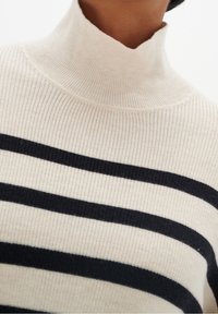 Turtlenecktröja i ribbat tyg, krämfärgad med svarta horisontella ränder. Har en åtsittande hals och en strukturerad yta.