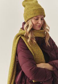 Cappello e sciarpa a costine color giallo senape abbinati a una giacca imbottita color bordeaux. La sciarpa è avvolta intorno al collo, mettendo in risalto la sua morbidezza.