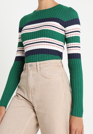 Kvinde iført en grøn ribstrikket sweater med hvide, marineblå og pink vandrette striber, kombineret med beige højtaljede bukser.