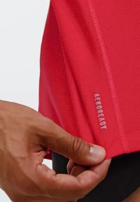 Camiseta deportiva roja con tela texturizada, con el logo "AEROREADY" en el costado. La mano tira ligeramente del dobladillo hacia arriba.