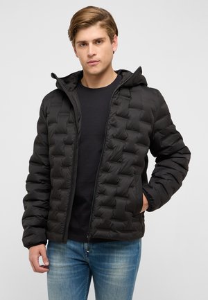 Junger Mann, der eine schwarze gesteppte Kapuzenjacke über einem schwarzen Hemd und blauen Jeans trägt, steht mit einer Hand in der Tasche.