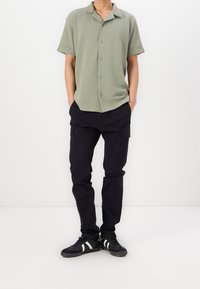 Zielona koszula z krótkim rękawem i kołnierzykiem, czarne spodnie bojówki z bocznymi kieszeniami oraz czarne sneakersy z białymi paskami, na jednolitym tle.