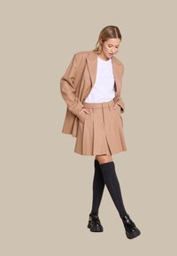 Lys brun skreddersydd blazer over en hvit t-skjorte, kombinert med en matchende plisserte skjørt. Tilbehør inkluderer svarte knelang sokker og platåsko.