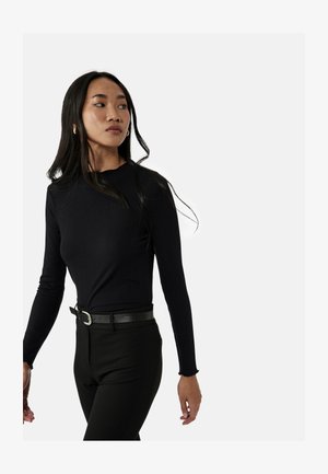 Ung kvinde med langt mørkt hår, iført en sort ribbet langærmet top og sorte bukser med sort bælte, ser mod venstre.