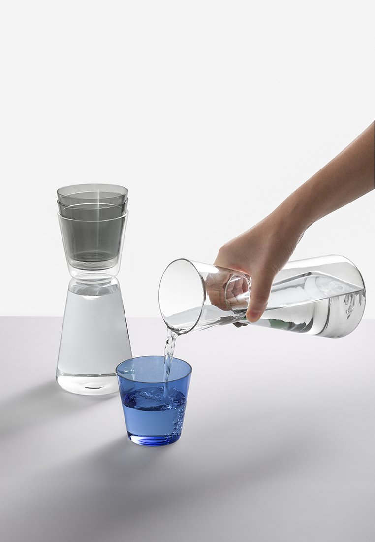 Przezroczysta szklana karafka wylewająca wodę do niebieskiego kubka. Elegancki, cylindryczny kształt o przezroczystym wykończeniu i gładkiej fakturze.
