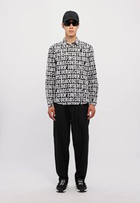 Graphic black and white long-sleeve shirt featuring repeating text pattern, paired with black wide-leg pants and black sneakers; wearing sunglasses and cap.