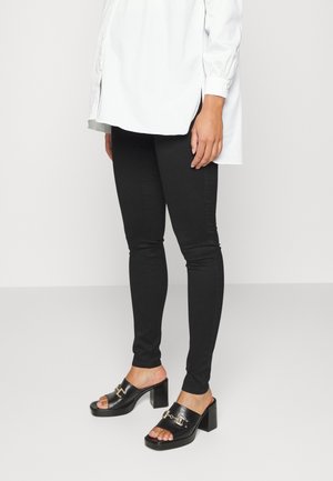 Jeans ajustados negros combinados con una camisa larga blanca. La modelo lleva sandalias de tacón negro tipo slip-on con una hebilla decorativa. Textura de tela suave.