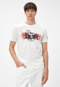 Camiseta de algodón blanca con un estampado gráfico de figuras históricas en negro con acentos rojos; diseño de manga corta y cuello redondo.