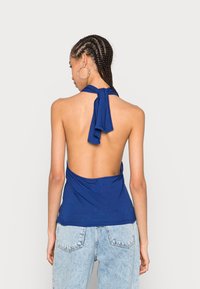 Vista traseira de um top halter azul com costas baixas e tecido drapeado, apresentando um fecho de pescoço amarrado e textura suave. Combinado com jeans claros.