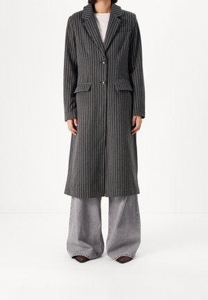 Femme portant un long manteau gris foncé à fines rayures sur un jean gris clair à jambes larges et des chaussures marron pointues sur un fond blanc.