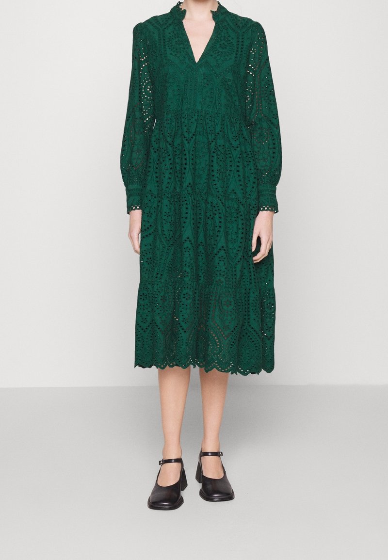 Kobieta ubrana w zieloną długą sukienkę midi z haftem i falbaniastym dołem, w czarnych butach Mary Jane, stojąca na jednolitym tle.