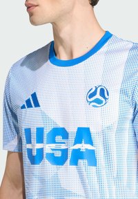 Bílý sportovní triko s modrými gradientními tečkami, s nápisem "USA" v tučných modrých písmenech, kulatým modrým límcem a logem Adidas.