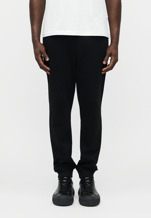 Mand iført sorte bukser, sorte sneakers og en hvid skjorte, stående på en neutral baggrund med hænderne afslappet langs siderne.