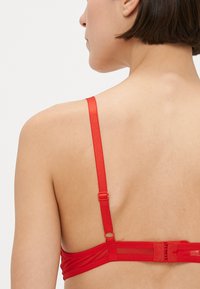 Rdeč nedrček iz gladkega blaga, z nastavljivimi naramnicami in zaponko za zapenjanje. Ima enobarvno zasnovo brez vzorcev ali okraskov.