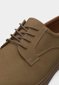 Καφέ υφασμάτινο casual παπούτσι με στρογγυλή μύτη, τέσσερις τρύπες για κορδόνια με αντίστοιχα καφέ κορδόνια και ορατή ραφή στο επάνω μέρος και τη γλώσσα.