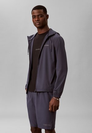 Ung mand iført en mørkegrå hættetrøje, matchende shorts og en sort Calvin Klein t-shirt, står mod en ensfarvet grå baggrund.