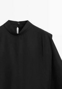 Chemise noire à col montant avec fermeture par bouton dans le dos et détails de coutures minimalistes à l’avant et aux épaules.