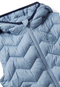 Světle modrá prošívaná bezrukávová bunda se zapínáním na zip a vyšitým logem blízko hrudi na bílém pozadí.