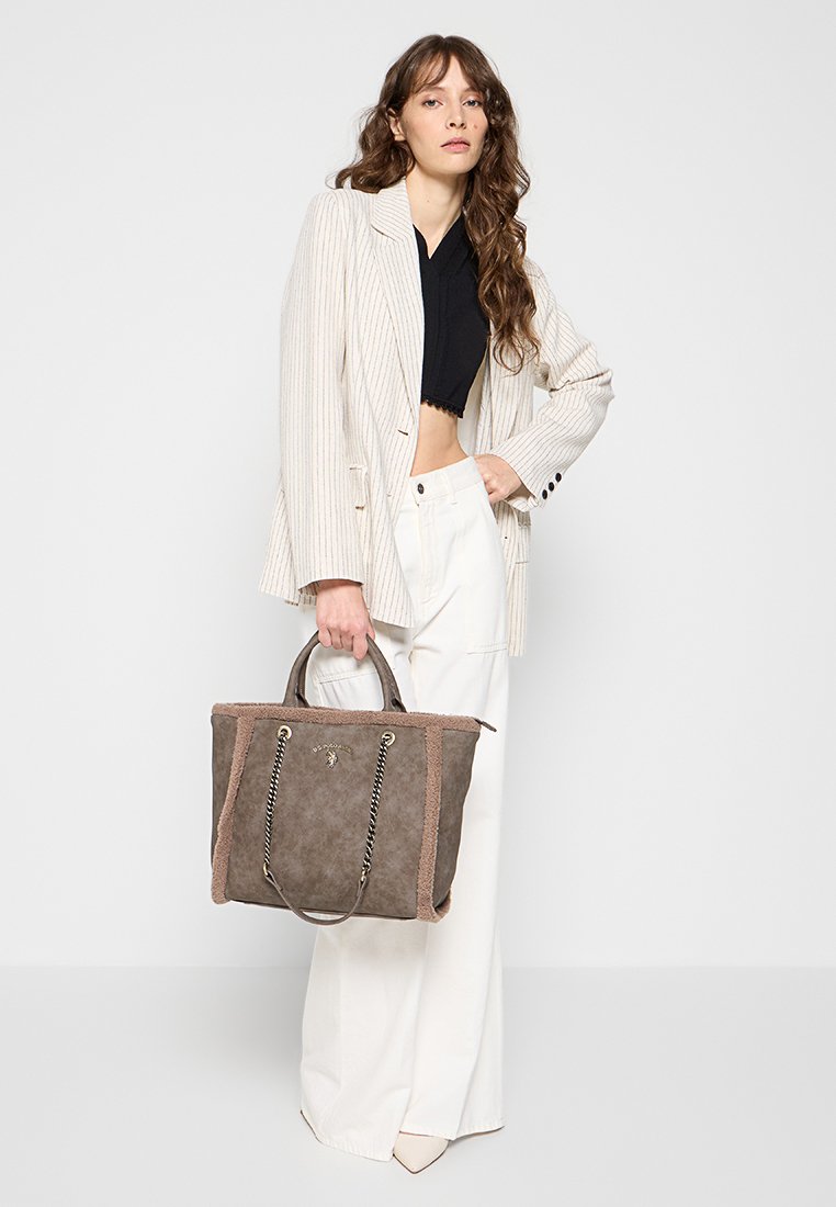 Sac fourre-tout gris et marron avec une surface texturée, des détails en chaîne et des poignées courtes, associé à un blazer rayé et un pantalon blanc.