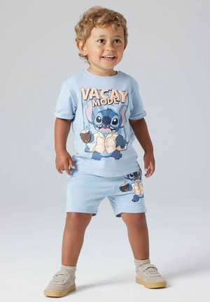 Toddler with curly hair wearing light blue "Vacay Mode" outfit featuring cartoon character holding drink, standing on plain background.