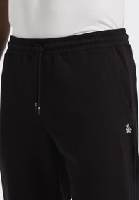 Zwarte sweatpants met een elastische tailleband, een trekkoord, zijzakken en een klein wit pinguïnlogo op het lagere linkerbeen. Zachte stoftextuur.