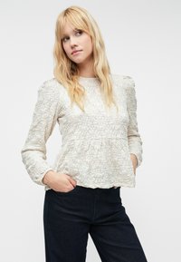 Blusa longa de manga comprida em bege claro com padrão floral texturizado, corpete franzido e decote redondo. Combinada com calças de ganga escuras.