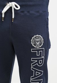 Mörkblå träningsbyxor i mjukt tyg, med vit snörning, broderad logotyp och "FRAN" textdetalj i vitt.