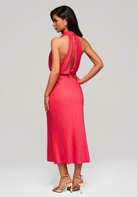 Robe en satin rouge avec un col halter et un dos nu, présentant un design drapé et une longueur mi-mollet. Associée à des sandales à talons hauts dorées.