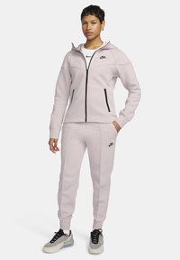 Chándal rosa claro de Nike que incluye una chaqueta con capucha y cremallera con detalles en negro y pantalones jogger a juego, confeccionado con un material suave y liso.