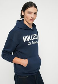 Námořnická modrá mikina s kapucí se klokaní kapsou a bílým nápisem "HOLLISTER So. California", vyrobená z měkké látky s hladkou texturou.