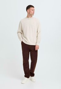 Beige sweatshirt with ribbed texture, round neckline, and a subtle logo; paired with dark brown corduroy pants and white sneakers.