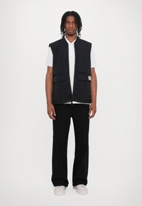 Black sleeveless vest with front pockets and textured accents, worn over a white polo shirt and paired with black pants.