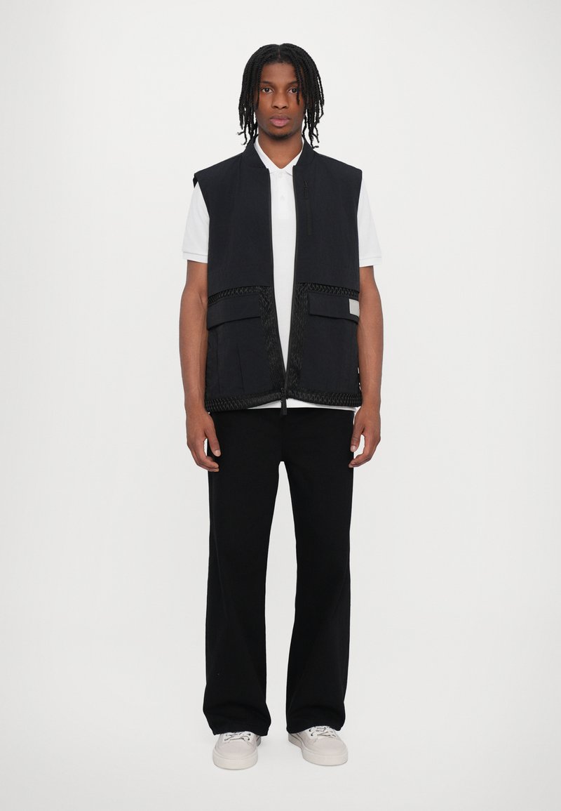 Black sleeveless vest with front pockets and textured accents, worn over a white polo shirt and paired with black pants.
