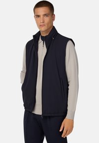 Marinblå väst med hög krage, dragkedja fram och sidofickor, tillverkad av ett smidigt, lätt material, i kombination med en ljusgrå långärmad topp.