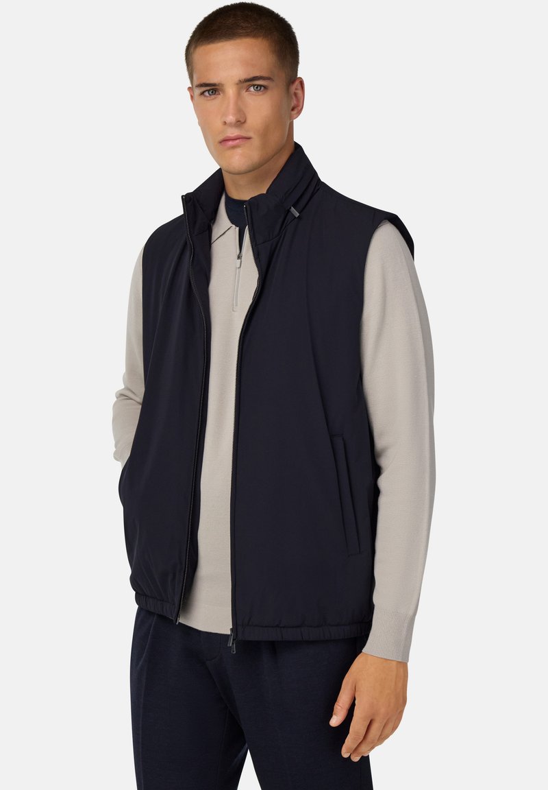 Marinblå väst med hög krage, dragkedja fram och sidofickor, tillverkad av ett smidigt, lätt material, i kombination med en ljusgrå långärmad topp.