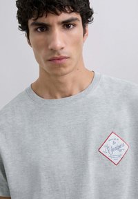Camiseta de algodón gris con un parche en forma de diamante con borde rojo y texto azul. Diseño de mangas cortas y cuello redondo.