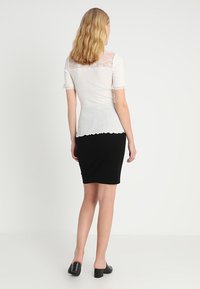 Top blanco con ribete de encaje y mangas cortas, tejido texturizado, corte ajustado; combinado con una falda negra hasta la rodilla y zapatos negros de tacón grueso.