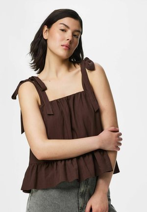 Jeune femme aux cheveux bruns mi-longs porte un haut marron sans manches avec des bretelles nouées et un pantalon gris sur un fond uni.
