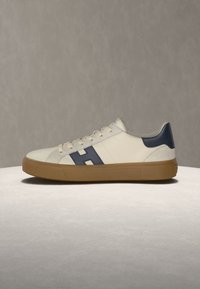 Λευκό sneaker χαμηλού τύπου με σόλα από καουτσούκ, μπλε ταμπελάκι στη φτέρνα και μπλε λογότυπο "H" στο πλάι, τοποθετημένο σε ανοιχτή επιφάνεια με φόντο γκρι.