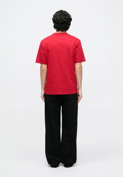 Κόκκινο βαμβακερό T-shirt με λαιμόκοψη και κοντά μανίκια, συνδυασμένο με μαύρο παντελόνι φαρδιάς γραμμής, από πίσω σε απλό φόντο.