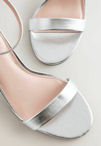 Sandalias metálicas plateadas con una textura suave, que presentan una amplia correa delantera y un diseño minimalista, sobre una plantilla de color rosa claro.