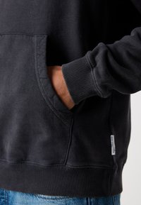 Hånd hvilende inde i den forreste lomme på en mørk sweatshirt med ribbede manchetter og kant, båret over et par blå jeans.