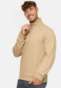 Pull-over beige à zip quart, en tissu doux, avec un col montant, des poignets côtelés et un petit logo sur la poitrine.