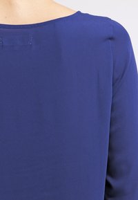 Blå bluse med en glat overflade, rund halsudskæring og lange ærmer, med diskrete syninger langs ryggen.