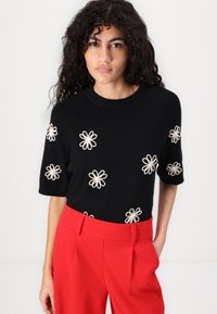 Pull en tricot noir à manches courtes, orné de marguerites blanches brodées. Associé à un pantalon rouge taille haute. Encolure ronde simple.