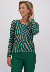 Langarmtop in een tan basis met groene en marineblauwe abstracte strepen. Heeft een V-hals en een getailleerd silhouet. Gecombineerd met gestructureerde groene broek.