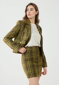 Blazer en tweed tissé jaune et noir avec des épaules structurées, associé à une jupe ajustée assortie. Surface texturée avec des accents dorés.