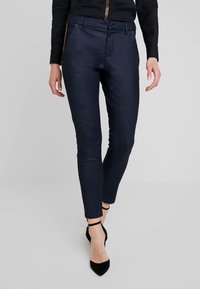 Pantalones ajustados de un oscuro tono navy con una textura suave, que presentan bolsillos laterales con cremallera y una cintura de altura media, combinados con tacones negros de punta afilada.